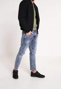 Schwarze Bomberjacke über einem dunkelgrünen Shirt, kombiniert mit hellblauen, ausgewaschenen Denim-Jeans und schwarzen Sneakers.