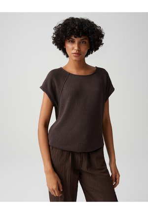 Femme aux cheveux courts et bouclés portant un haut marron ample à manches courtes et un pantalon marron assorti, debout devant un fond clair et uni.