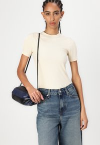 Μπεζ ριμπ μπλουζάκι κοντό μανίκι σε συνδυασμό με ψηλόμεσα μπλε τζιν, κρατώντας μια μικρή μαύρη και ναυτική τσάντα με λουρί ώμου.