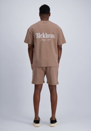 Man staat met zijn rug naar de kijker, draagt beige shorts en een bijpassend oversized T-shirt met de tekst "Blckbnns Design Dept" in wit op de achterkant.