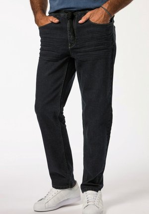 Homme portant un pantalon jogger en jean noir avec taille à cordon et des baskets blanches, les mains dans les poches, sur un fond uni.