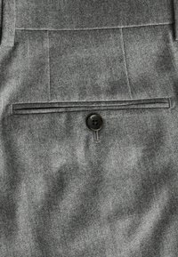 Una tela gris con una textura fina presenta un solo botón en un bolsillo de solapa. El bolsillo tiene un detalle de costura horizontal por encima.