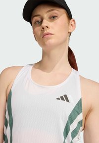Bijela sportska majica bez rukava s zelenim dijagonalnim prugama, teksturirana tkanina i crni logo. Nosena s crnom kapom, jednostavnog dizajna.