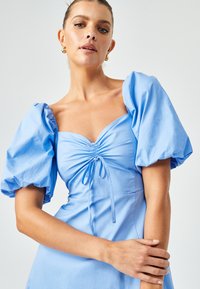 Heledsinine kleit puffvarrukatega, tiheda rinnaosa ja kogutud esiosaga, millel on nööri detail. Sile ja struktureeritud kangas.