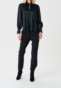 Top in satin nero con scollatura a chiave e maniche a sbuffo, abbinato a pantaloni neri a sigaretta con polsini arricciati e stivali con tacco a spillo.