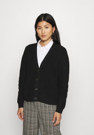 Zwarte gebreide cardigan met een V-hals, vijf knopen, geribbelde textuur en lange mouwen, gedragen over een witte blouse met kraag en geruite broek.