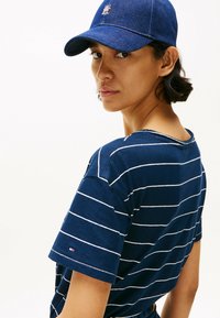 Camiseta de rayas azul marino con mangas cortas, que presenta rayas blancas. Una gorra de mezclilla complementa el atuendo. Detalle del logo en la gorra y la camiseta.
