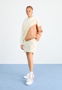 Sweatshirt à capuche avec un design en blocs de couleurs crème et marron ; associé à une jupe cargo beige et des baskets blanches avec des accents rose.