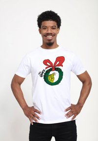 Biała bawełniana koszulka z zielonym Grinchem w okrągłym wieńcu z czerwonymi akcentami i dużą czerwoną kokardą. Napis brzmi "Grinch".