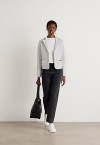 Gray blazer with black accents, white shirt, black jeans, and white sneakers. Model holds a black tote bag. Smooth fabric, tailored fit.