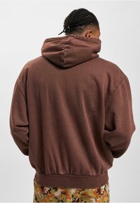 Sudadera marrón con un acabado texturizado y capucha ajustable, que presenta una cintura acanalada. Combinada con pantalones de patrón de camuflaje.