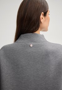 Grijze pullover met een hoge kraag, voorzien van een gladde textuur en een decoratieve ronde knop aan de achterkant. Eenvoudig ontwerp met minimale details.