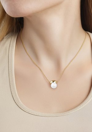 Guld kæde halskæde med en enkelt stor hvid perlevedhæng båret på en kvindes hals over en beige top.