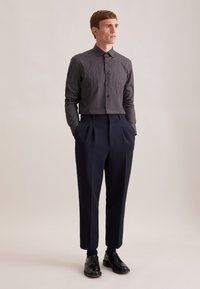 Marineblauwe broek met plooien, gecombineerd met een donker gestreept overhemd met knopen. Materiaal lijkt glad; het overhemd heeft een geometrisch ontwerp. Zwarte schoenen maken de look compleet.