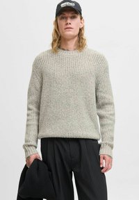 Suéter de punto texturizado en gris claro con patrón de canalé, escote redondo y mangas largas, combinado con pantalones de pinstripe negros y sujetando una prenda negra.