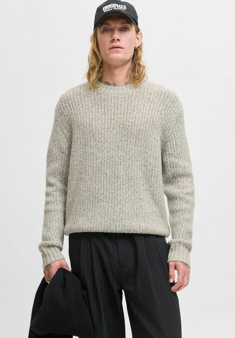 Hellgrauer strukturierter Strickpullover mit Rippenmuster, Rundhalsausschnitt und langen Ärmeln, kombiniert mit schwarzen Nadelstreifen-Hosen und hält ein schwarzes Kleidungsstück.