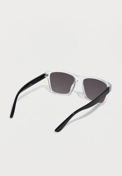 Γυαλιά ηλίου με διαφανή ορθογώνια πλαίσια και μαύρους βραχίονες, φακούς με σκουρόχρωμη απόχρωση, ανοιγμένα και τοποθετημένα σε λευκή επιφάνεια.