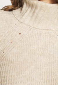 Vaaleanbeige poolopaita, jossa on ribbikuvio, hienovarainen vinokudontakuvio ja pieniä reikiä lisäyksinä. Pehmeä ja mukava kangas.