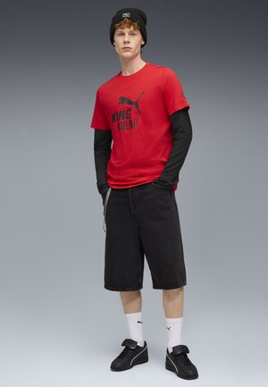 Red T-shirt with black "KING MILAN" print, worn over a black long-sleeve shirt, paired with black denim shorts and black sneakers.