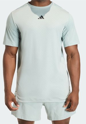 Mężczyzna w jasnoszarej sportowej koszulce z krótkim rękawem z czarnym logo Adidas na piersi i pasujących szortach, pokazany od szyi do połowy uda.