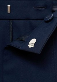 Tissu bleu marine avec une texture subtile, présentant une fermeture éclair visible, un bouton et un détail en clip argenté sur le bord du vêtement.