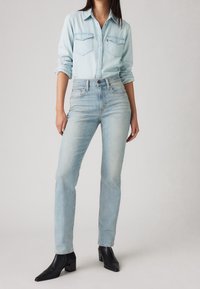 Light blue denim jeans with a high waist, straight fit, and subtle fading; paired with a light blue button-up shirt and black heeled boots.