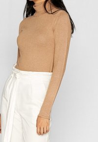 Top de manga larga y cuello alto en tejido de punto color beige claro, combinado con pantalones blancos de tiro alto que presentan un detalle de cinturón atado.