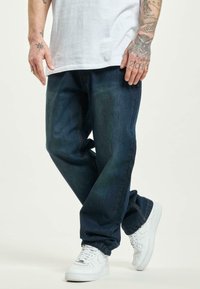 Pantalones de mezclilla azul oscuro de corte holgado, con costuras naranjas, puños ajustados y una textura suave, combinados con zapatillas blancas.