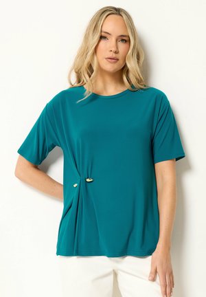 Femme aux cheveux blonds portant un haut bleu sarcelle à manches courtes avec des accents dorés et un pantalon blanc, debout sur un fond blanc.