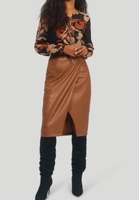 Kvinna i en brun blommig blus, brun fusk läder midi kjol med framficka, och svarta knähöga klackstövlar, stående med händerna ihopknäppta.