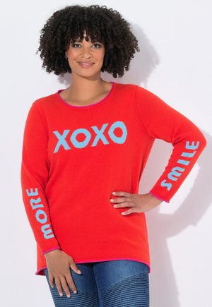 Vrouw met krullend haar die een felrode trui draagt met blauwe tekst "XOXO" op de voorkant, "MORE" op de linkermouw en "SMILE" op de rechtermouw.