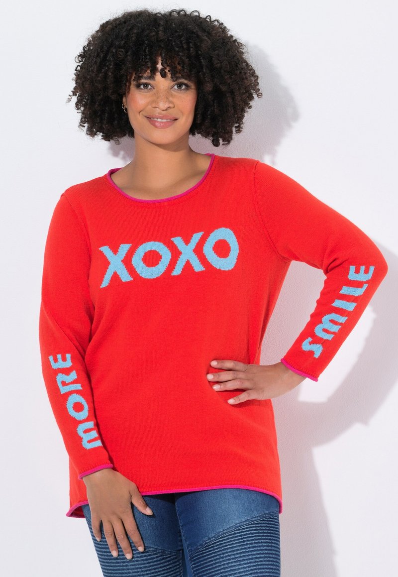 Femme avec des cheveux bouclés portant un pull rouge vif avec du texte bleu "XOXO" sur le devant, "MORE" sur la manche gauche et "SMILE" sur la manche droite.