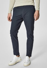 Navy blue slim-fit trousers made of a textured fabric, featuring a zip fly, belt loops, and front pockets, paired with white sneakers.