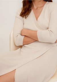 Femme assise les bras croisés, portant une robe portefeuille crème à manches longues, côtelée, avec une fente latérale.