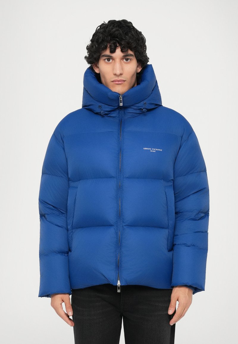 Plava podstavljena puffer jakna s visokim ovratnikom, zatvaranjem na zip i detaljem s logotipom na prsima. Ima oversized siluetu i elastične rukave.