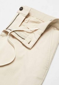 Beige Hose mit Reißverschluss, Knopfverschluss und Kordelzug. Der Stoff ist glatt mit einem leicht strukturierten Bund und Seitennähten.