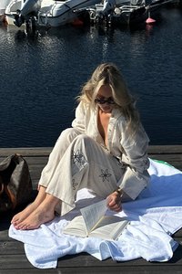 En person sitter på en vit handduk vid en brygga, iklädd en ljusbeige outfit med svart blomsterbroderi, och läser en öppen bok.