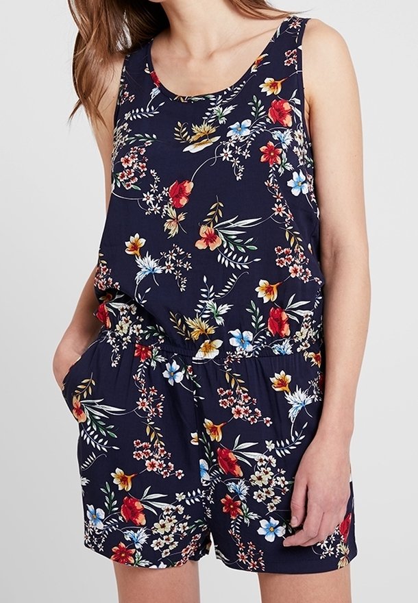 Combinaison sans manches bleu marine à fleurs avec taille élastique, présentant des fleurs rouges, jaunes, blanches et bleues, portée par une personne avec la main dans la poche.