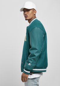 Teal varsityjack met leren mouwen, witte afwerking en voorop letters. Model draagt een witte pet en lichte spijkerbroek, waarbij de zijaanzicht wordt getoond.