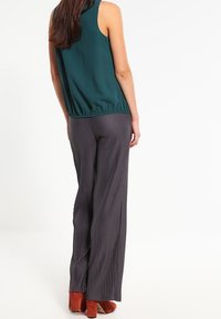Blouse sans manches bleu sarcelle avec taille froncée, associée à un pantalon gris à rayures verticales longues. Les chaussures sont brun-orange, avec une finition lisse.