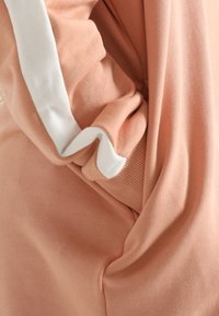 Světle broskvová mikina s bílým akcentem na rukávu; má měkkou texturu a volný střih, ukazuje nařasenou látku na manžetě.