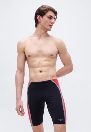 Nuori mies, jolla on lyhyet ruskeat hiukset ja joka on pukeutunut mustiin, punaisiin ja harmaisiin polvipitkiin uimalakkeihin, seisoo yksinkertaisen vaalean taustan edessä.