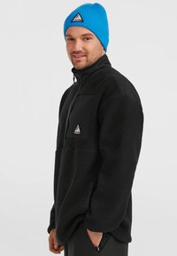Pull noir en polaire avec un col zippé, surface texturée, et une poche sur la poitrine. Porté avec un bonnet en tricot bleu présentant un logo triangulaire.