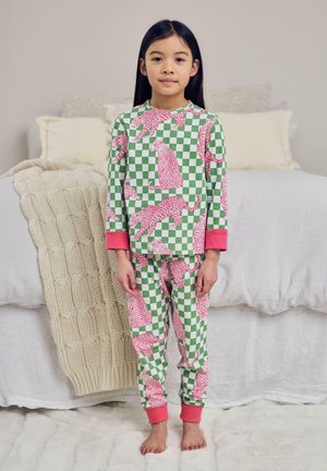 Junges Mädchen steht barfuß im Schlafzimmer und trägt grüne und weiße karierte Schlafanzüge mit pinken Leopardendrucken und pinken Bündchen.
