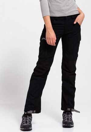 Personne portant un pantalon cargo noir et des chaussures de randonnée grises, debout avec une main dans la poche et l'autre détendue.