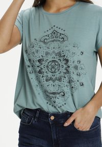 Světle tyrkysové tričko s detailním černým mandala grafickým designem ve středu. Krátké rukávy, volný střih, s měkkou texturou látky.