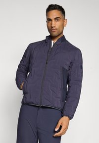 Veste rembourrée bleu marine, avec fermeture éclair sur le devant, poignets côtelés et accents en mailles. Présente une texture lisse et un design légèrement rembourré.