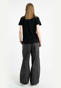 Osoba z prostymi, jasnobrązowymi włosami, ubrana w czarną koszulkę i szerokie, ciemnoszare spodnie, stojąca tyłem na białym tle.