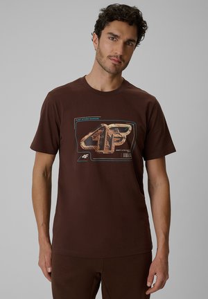 Mężczyzna z kręconymi włosami w brązowej koszulce z dużym logo 4F i graficznym napisem, stojący na tle jednolitego szarego tła.