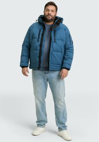 Modra podložena jakna s črnimi poudarki, z žepi na zadrgo in nastavljivo kapuco, v kombinaciji s svetlo modrimi kavbojkami in belimi teniskami.